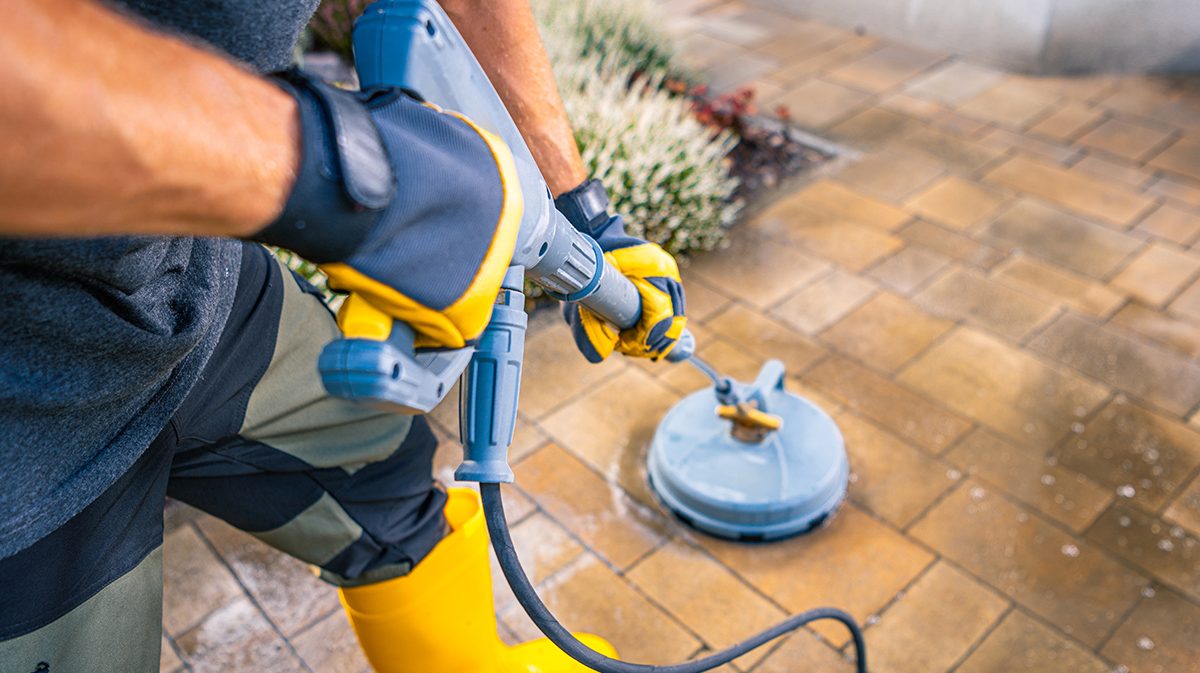 A person in yellow boots and gloves operates a pressure washer on a stone patio, effectively cleaning the surface in a residential backyard setting.